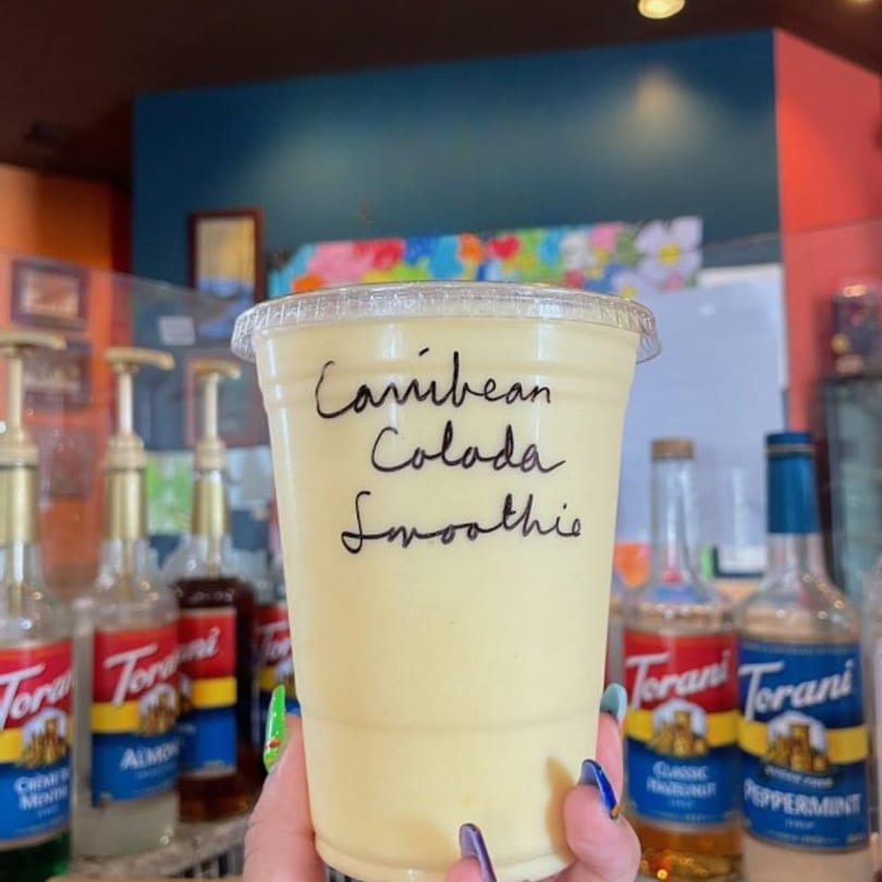 Hand holding a Caribbean Colada Smoothie from Grouchy John's Coffee, with syrup bottles in the background.