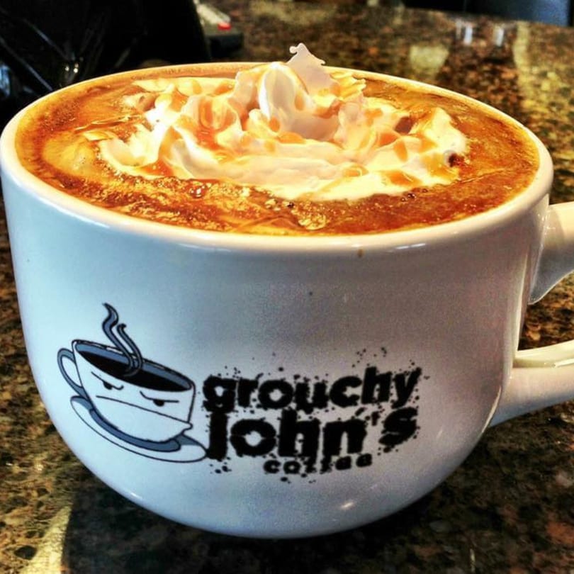 A Grouchy John's Coffee mug filled with a caramel latte topped with whipped cream and drizzle.